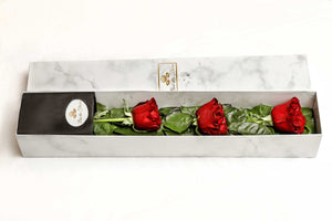 A close-up image of a flower arrangement from Vancouver's Box Des Fleurs, called "Mousquetaire" (Three Musketeers). The arrangement features three long-stemmed red roses in a triangular shape with lush green leaves in a white marble box. The minimalist and elegant design of the arrangement makes it perfect for any occasion, from romantic gestures to expressing appreciation.