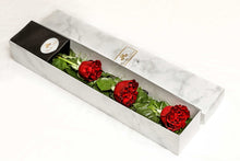 A close-up image of a flower arrangement from Vancouver's Box Des Fleurs, called "Mousquetaire" (Three Musketeers). The arrangement features three long-stemmed red roses in a triangular shape with lush green leaves in a white marble box. The minimalist and elegant design of the arrangement makes it perfect for any occasion, from romantic gestures to expressing appreciation.