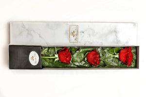 A close-up image of a flower arrangement from Vancouver's Box Des Fleurs, called "Mousquetaire" (Three Musketeers). The arrangement features three long-stemmed red roses in a triangular shape with lush green leaves in a white marble box. The minimalist and elegant design of the arrangement makes it perfect for any occasion, from romantic gestures to expressing appreciation.