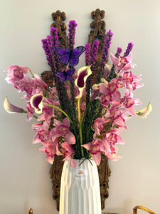 A close-up image of a flower arrangement from Vancouver's Box Des Fleurs, called "Jardin de Papillons" (Butterfly Garden). The arrangement features pink, white, purple, yellow, and green seasonally available flowers, including orchids and other delicate blooms, and green leaves in a round glass vase. Two butterfly-shaped ornaments are perched on branches inside the vase, adding a whimsical touch. Perfect as a gift or to brighten up any room.