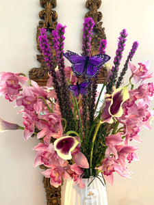 A close-up image of a flower arrangement from Vancouver's Box Des Fleurs, called "Jardin de Papillons" (Butterfly Garden). The arrangement features pink, white, purple, yellow, and green seasonally available flowers, including orchids and other delicate blooms, and green leaves in a round glass vase. Two butterfly-shaped ornaments are perched on branches inside the vase, adding a whimsical touch. Perfect as a gift or to brighten up any room.