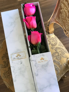 A close-up image of a flower arrangement from Vancouver's Box Des Fleurs, called "Mousquetaire" (Three Musketeers). The arrangement features three long-stemmed magenta pink roses in a triangular shape with lush green leaves in a white marble box. The minimalist and elegant design of the arrangement makes it perfect for any occasion, from romantic gestures to expressing appreciation.
